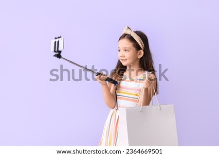 Cute little girl with shopping bag taking selfie on lilac background
