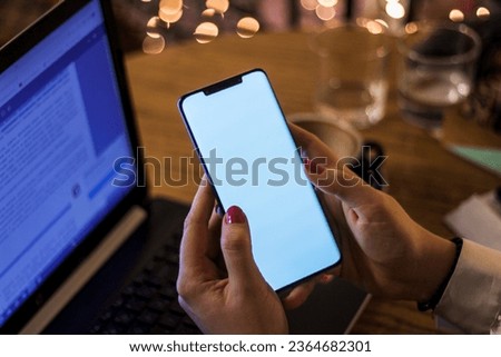 Young woman holding a smartphone with blank white screen while working on a laptop in a coffee shop. Horizontal photo with copy space for text, logo or add