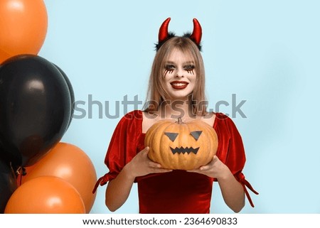 Young woman dressed for Halloween as devil with pumpkin and balloons on blue background