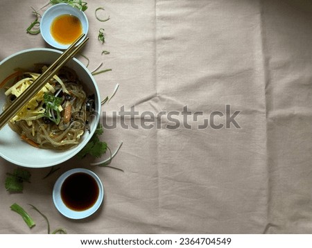 japchae is a savory dish of stir-fried glass noodles and vegetables that is popular in Korean cuisine.Pictured with condiment on a light brown condiments.Horizontal image. copy space. Negative space.