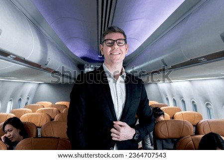 Portrait of happy smiling businessman in black suit, standing on aisle inside airplane, male passenger traveling on business trip by aircraft, businesspeople traveling with airline transportation. Royalty-Free Stock Photo #2364707543