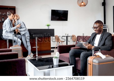 Happy smiling African businessman with luggage sitting on sofa in airport business lounge, busy male passenger working on tablet at waiting area of airport, waiting flight at airport departure lounge Royalty-Free Stock Photo #2364708119