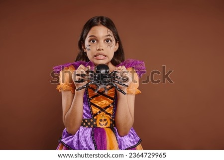 spooky girl in Halloween costume holding fake spider and grimacing on brown backdrop, October 31