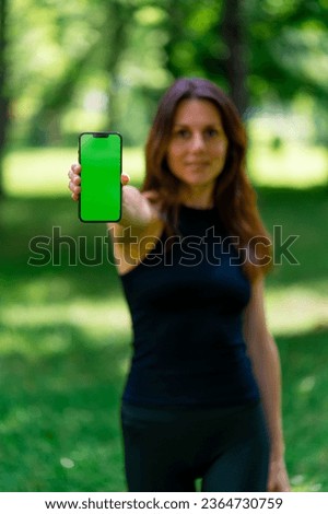 Active beautiful yogi woman during a walk in the park before or after training stands holding a smartphone with green screen in her hands
