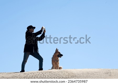 Woman with dog on sand dune under blue sky, taking photographs with a smartphone. Copy space