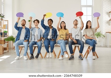 Group of happy young diverse multiracial people holding up colorful paper mockup speech bubbles while sitting in row on chairs in new modern office or rented apartment. Opinion survey concept