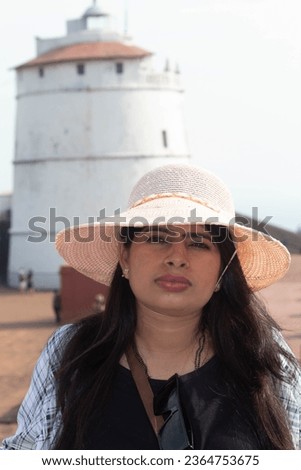 Portrait of stylish female traveler in sun hat. She is looking at camera while standing in front of ancient lighthouse