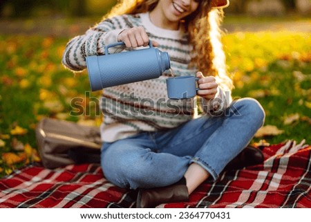 Cute woman in a hat and a stylish sweater sits on a blanket in an autumn park and drinks a hot drink from a thermos. A tourist woman enjoys the autumn weather in the park. Vacation, weekend concept.