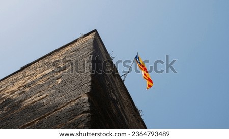 Catalan flag waving in the blue sky on an ancient medieval tower. Red and yellow striped flag on historic stone building.