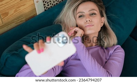 Young beautiful blonde woman 20 years old in casual clothes, purple home suit, taking first person selfie shot on mobile phone, sitting on sofa indoors, relaxing at home in own room of apartment