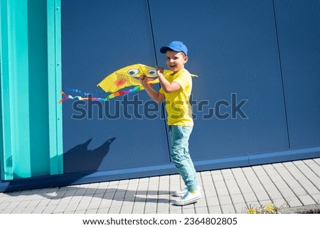 Little seven year old boy in a yellow T-shirt and blue baseball cap, white sneakers playing with his yellow kite emoji outside against a blue wall Royalty-Free Stock Photo #2364802805