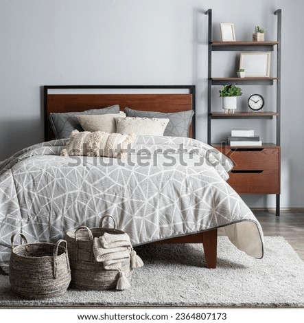 Cozy Modern Bedroom with Light Gray Wall, Featuring a Wooden Bed with Gray Duvet, Decorative Baskets, and a Wooden Bookshelf with Home Decor Items, front view. Royalty-Free Stock Photo #2364807173