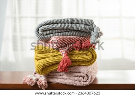 Neatly Stacked Knitted Blankets with Diverse Designs and Textures, Adorned with Tassels, Displayed on a Wooden Table in a Bright Room with Window Illumination in the Background Royalty-Free Stock Photo #2364807623