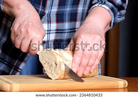 The hands of an elderly woman cut white wheat bread baguette with a knife in the kitchen at a brown table, without a face, close-up