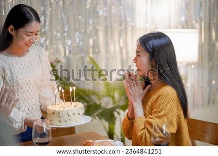 Cheerful friends enjoying home Birthday holiday party. Asian sister cheering drinking red wine celebrating with Birthday cake
