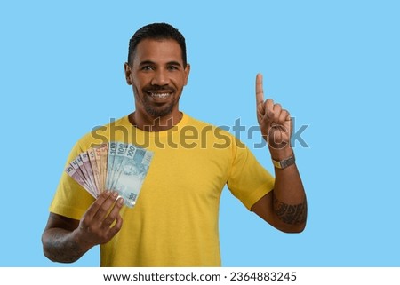 A man with Brazilian money in his hand points upwards with his finger to an empty space on the right-hand side of the picture, yellow clothes, blue background