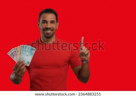 A man with Brazilian money in his hand points upwards with his finger to an empty space on the right-hand side of the image, wearing a red T-shirt, red background.