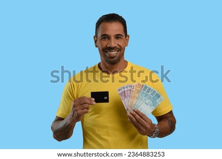 Man with a black credit card and Brazilian money in his hand, yellow clothes, blue background