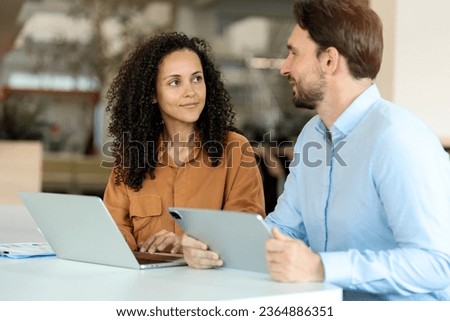 Beautiful curly African American female manager using laptop, sitting at desk, working together with hispanic bearded male colleague in modern office. Concept of successful career Royalty-Free Stock Photo #2364886351