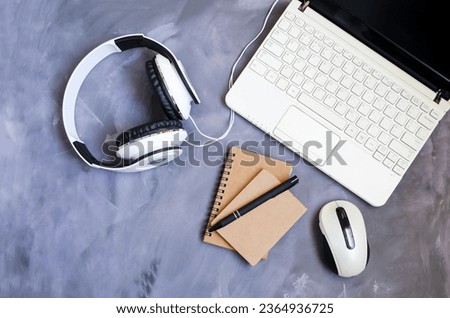 An open laptop stands on a table on a gray background, next to it lies a notepad, a pen, white headphones and a computer mouse.  Concept of office work, online training, webinars.  Flat laying.