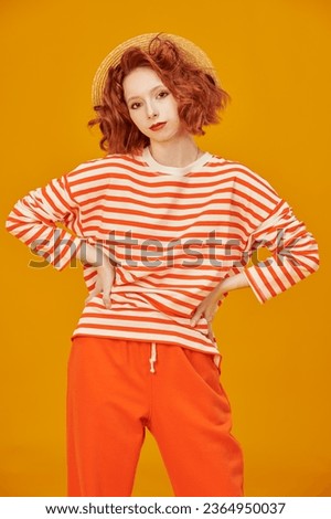 People's emotions. Beautiful bright girl with curly red hair, dressed in a straw hat and longsleeve with orange stripes posing on a yellow studio background. Summer fashion and beauty.