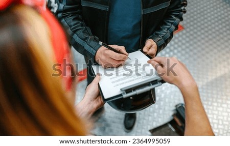 Unrecognizable female mechanic with red hair bandana holding a clipboard while biker man wearing leather jacket signing insurance policy to receipt his repaired motorcycle Royalty-Free Stock Photo #2364995059