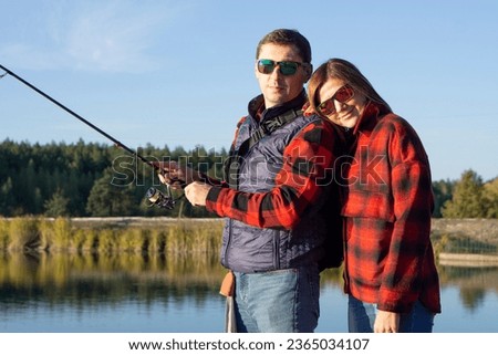 Fishing date. Man teaching his wife to fish, middle-aged family couple wearing matching red plaid shirts fishing together by lake, trying to catch fish, bonding while spending time in nature