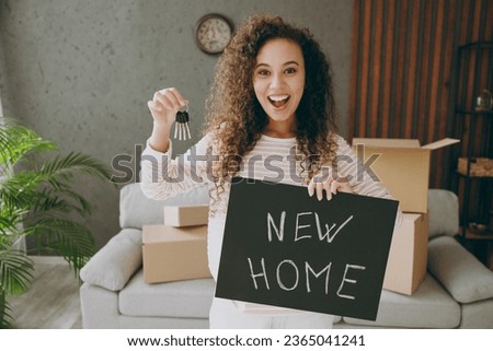 Young surprised woman wear casual clothes sits near grey sofa couch hold card sign with new home title text, keys stay at flat rest relax spend free spare time in living room indoor. Lounge concept Royalty-Free Stock Photo #2365041241