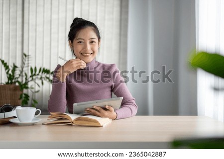 A portrait of a beautiful and smiling young Asian woman in casual clothes sits at a table in her living room with her digital tablet. People and technology concepts