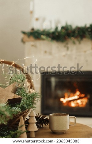 Cozy winter. Stylish cup of warm tea, basket with fir branches, wooden trees and star, pine cones on table against burning fireplace. Modern rustic eco friendly decor in scandinavian room