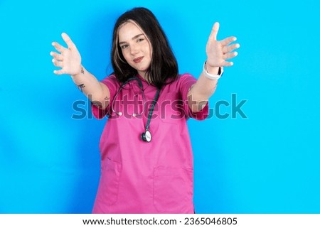 young beautiful doctor woman standing over blue background looking at the camera smiling with open arms for hug. Cheerful expression embracing happiness. Royalty-Free Stock Photo #2365046805