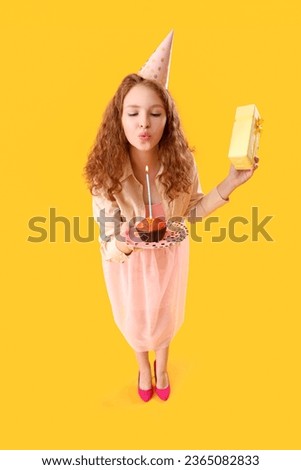 Beautiful woman with Birthday gift blowing out candle on cake against yellow background