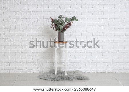 Vase with green eucalyptus branches on end table near white brick wall