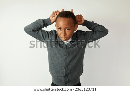 Funny african american male kid in school uniform having fun during break between lessons, making horns on his head with fingers, grimacing, frighting his mates, saying boo on white studio background