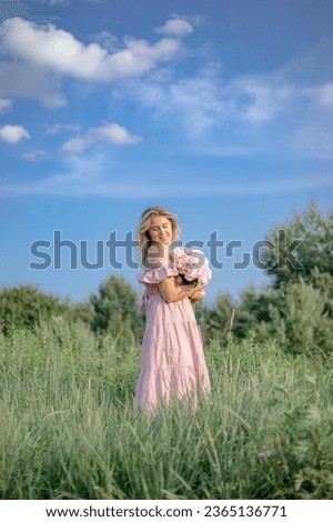 Vertical image. Beautiful woman in pink summer sundress with large bouquet of peonies stands among green grass against blue sky. Femininity, beauty. Positive emotions. Natural cosmetics, skin care. 