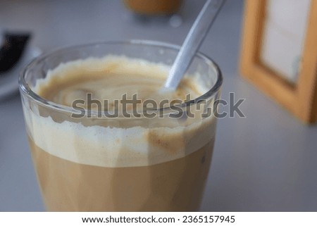 Picture of a coffee with milk in a glass and with a spoon inside. There is a very tasty foam on top. On the background, paper napkins on the table. Image taken in a cafeteria. Latte macchiato.