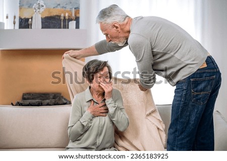 Loving supportive mature man take care of his ill and elderly wife, covering her with plaid while she sitting on couch at home, suffering from flu virus disease Royalty-Free Stock Photo #2365183925