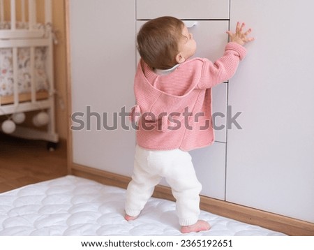 10 month old baby girl dressed in pink hoodie tries to stand on her feet and leans his hands on the childrens chest of drawers. Royalty-Free Stock Photo #2365192651
