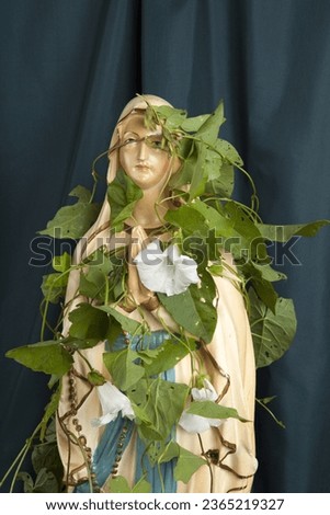 a statuette of the Virgin Mary covered with bindweed in front of a wavy green curtain. a metaphor for time and its poetry. Minimal still life photography