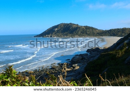 View of a beach from a hill. The waves are crashing on the beach and you can see another hill and the blue sky in the background. The picture was taking during the sunset in a winter day.