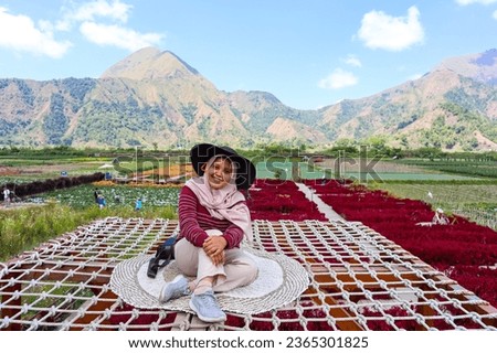 A woman with hijab and floppy hat smile and sit on selfie area in agriculture flower park with mountain in the background during vacation Royalty-Free Stock Photo #2365301825