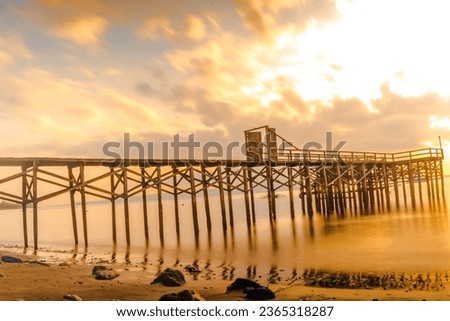 A wooden pier stretches out into a calm body of water, bathed in the golden light of sunset. The sky is ablaze with color, and the reflection of the pier in the water is stunning.