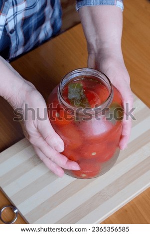 The hands of an elderly woman take out delicious homemade pickled tomatoes from a jar in the kitchen at a brown table, without a face, close-up