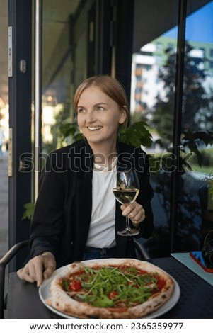 Young business woman drinks white wine from a glass of wine in a street cafe, looking at the camera. Wine tasting. Italian wine. Blurred light background.Italian pizza on the table. Copy space.