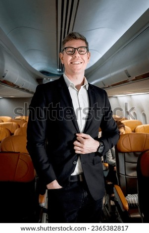 Portrait of happy smiling businessman in black suit, standing on aisle inside airplane, male passenger traveling on business trip by aircraft, businesspeople traveling with airline transportation. Royalty-Free Stock Photo #2365382817