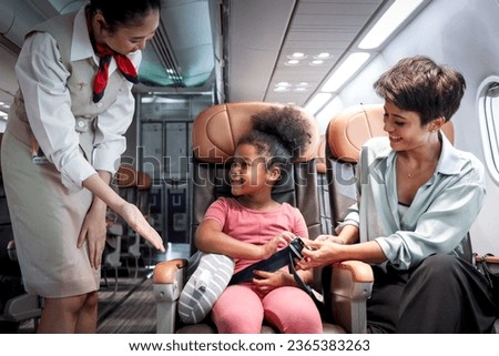 Friendly air hostess take care kid passenger on airplane, flight attendant teach curly hair African girl to use seat belt during sit in aircraft seat, child traveling by plane, airline transportation Royalty-Free Stock Photo #2365383263