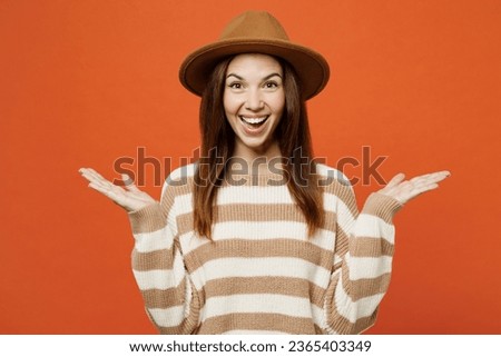 Young overjoyed excited fun cool woman she wearing striped sweater hat casual clothes look camera spread hands isolated on plain orange red color wall background studio portrait. Lifestyle concept