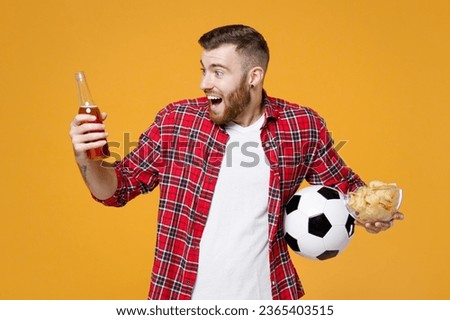 Surprised young man football fan in red shirt cheer up support favorite team with soccer ball bottle of beer bowl of chips isolated on yellow background studio portrait. People sport leisure concept