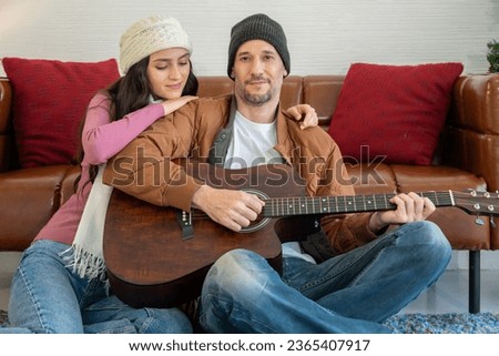 Couple lover in sweater enjoying great time in living room with guitar. Young man playing guitar and singing song together with girlfriend while sitting on floor together in front of sofa