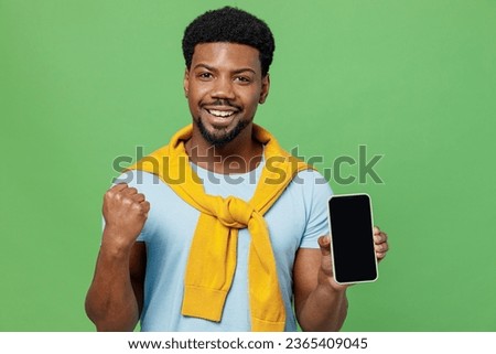 Young excited man of African American ethnicity 20s wear blue t-shirt hold in hand use mobile cell phone with blank screen workspace area do winner gesture isolated on plain green background studio.
